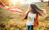 Little girl playing with a ring that has ribbons attached wearing a traveling band t-shirt that has a little cute car in a field of yellow flowers