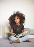 Little girl wearing a LouliiBot black t-shirt reading a book on the counch.