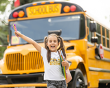 Little girl  wearing a traveling band t-shirt that has a little cute car in a field of yellow flowers holding markers in front of a school bus