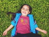 Little girl laying on grass laughing wearing a dark pink Flavor Fruit Day toddler t-shirt