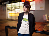 Funky Tee that shows a seventies funky green color print of the word Funky in the color white worn by a young women leaning against a bike rack wearing also a black jacket